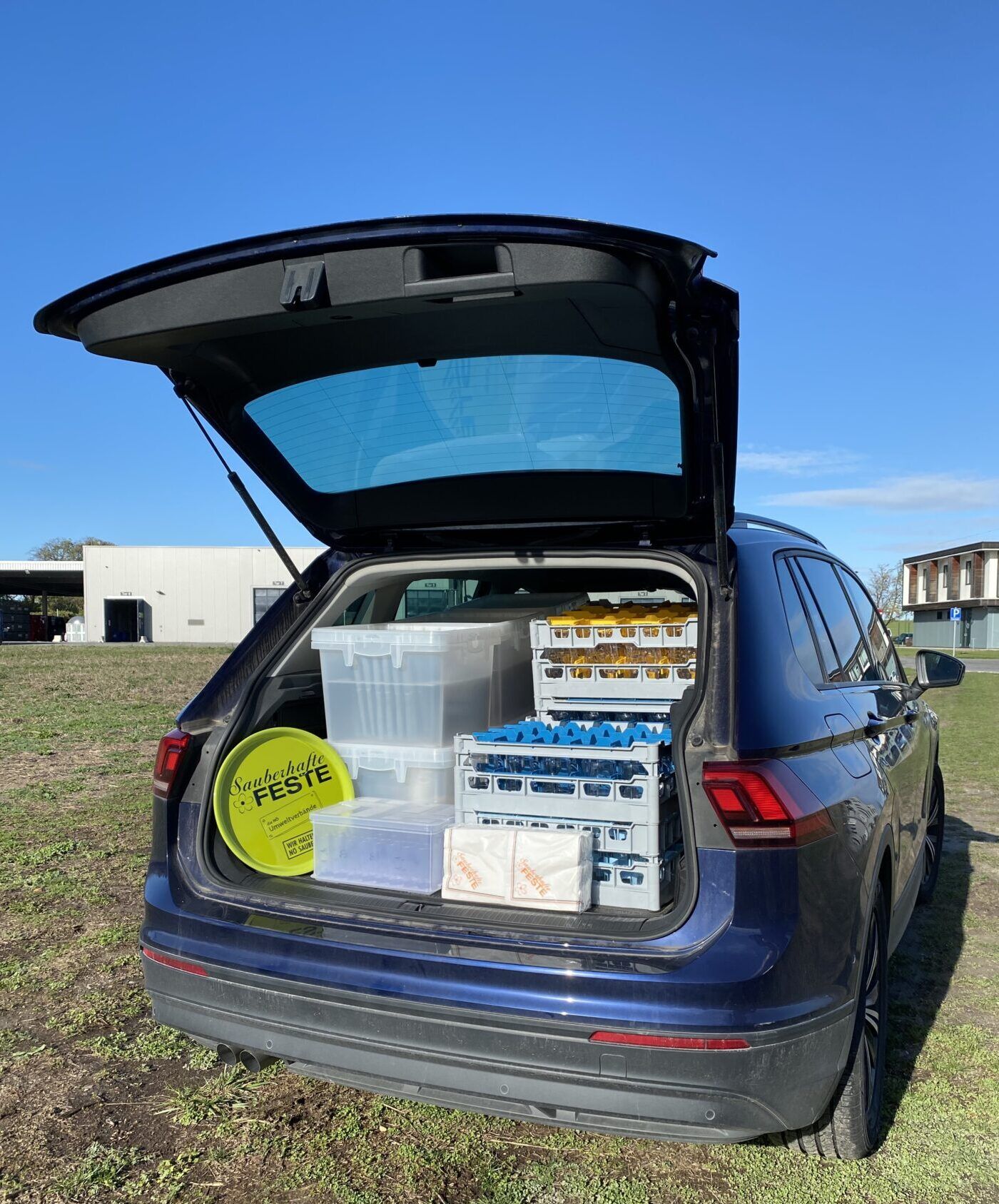 Ein Auto steht in der Wiese, der Kofferraumdeckel ist geöffnet. Im Kofferraum befindet sich das Festpaket To Go mit Gläsern, Geschirr, Besteck, Tablett und Servietten.