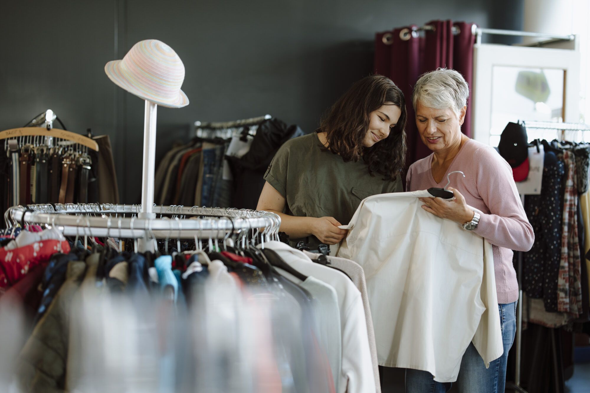 Eine Jugendliche mit schwarzen Haaren und eine ältere Dame mit grauen Haaren begutachten eine gebrauchte weiße Bluse im ReUse Shop. Vor ihnen ein Kleiderständer mit einer Vielzahl weiterer Kleidungsstücke