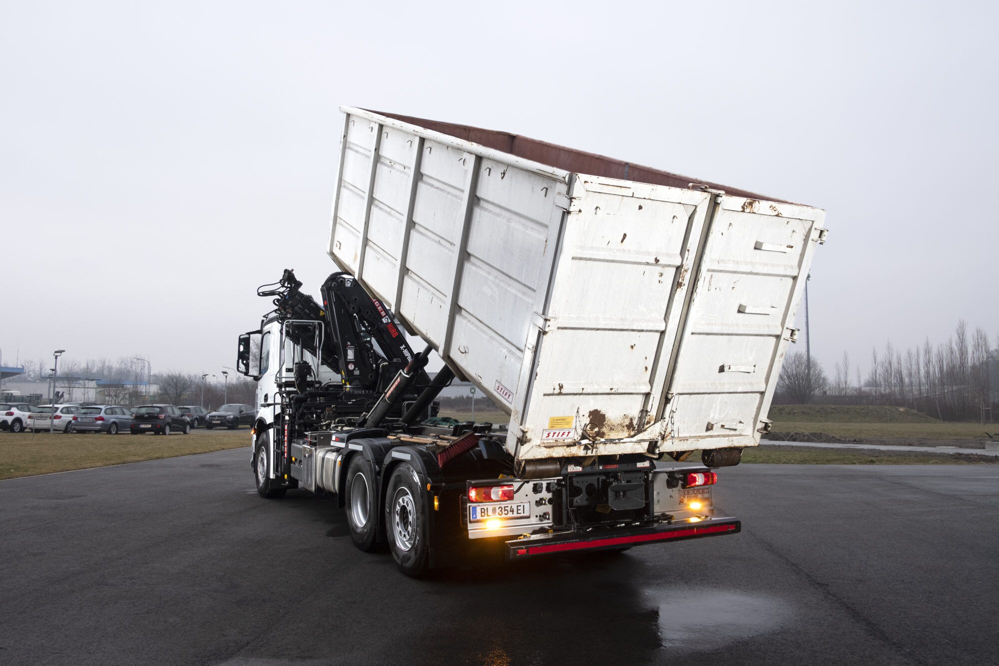 Ein LKW ist von hinten zu sehen, er ist dabei einen Container abzuladen, im Hintergrund sind Autos zu sehen