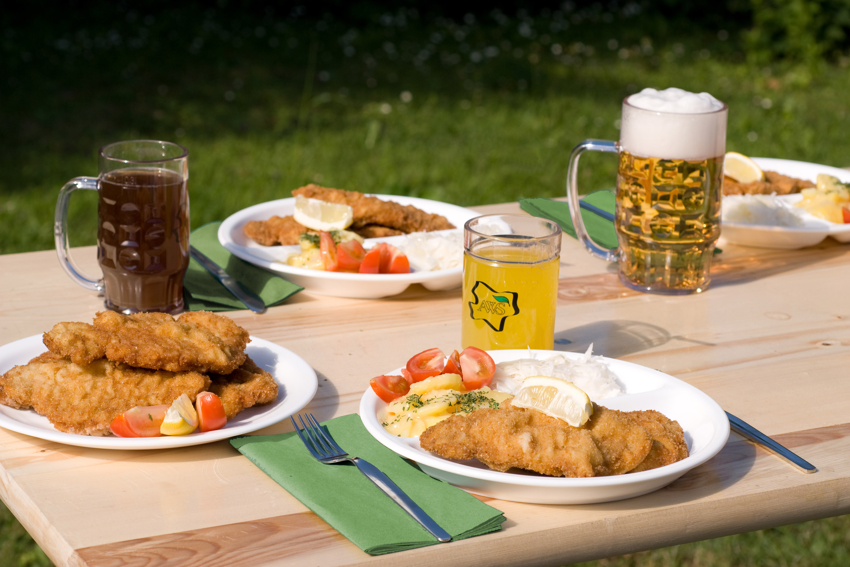 Gedeckter Tisch mit unserem AWS-Geschirr, 4 Teller mit Schnitzel und Beilagen, drei Gläser mit unterschiedlichen Limos und Bier. Es liegt Besteck am Tisch mit grünen Servietten.