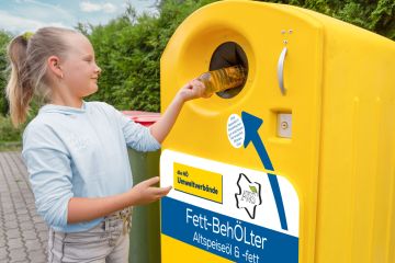 Blondes Mädchen wirft eine eine transparente Plastikflasche, in der sich Altspeiseöl befindet in das Einwurfloch eines gelben Sammelbehälters zum Sammeln von Altspeiseöl und -fett