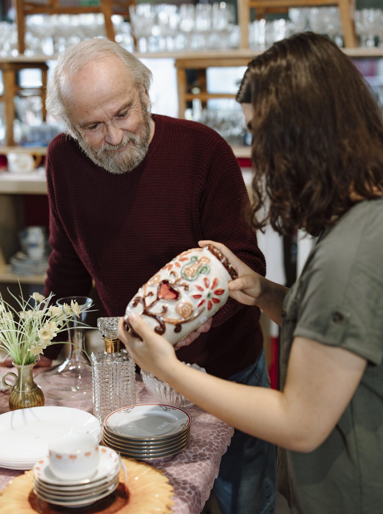 Re-Use Shop des AWS in Schwadorf älterer Herr mit weißen Haaren und Bart begutachtet gemeinsam mit jugendlichem Mädchen mit dunklen Haaren Re-Use Geschirr. Das Mädchen hält eine bunte Vase in den Händen und man sieht außerdem Teller, Tassen und weiteres Geschirr.