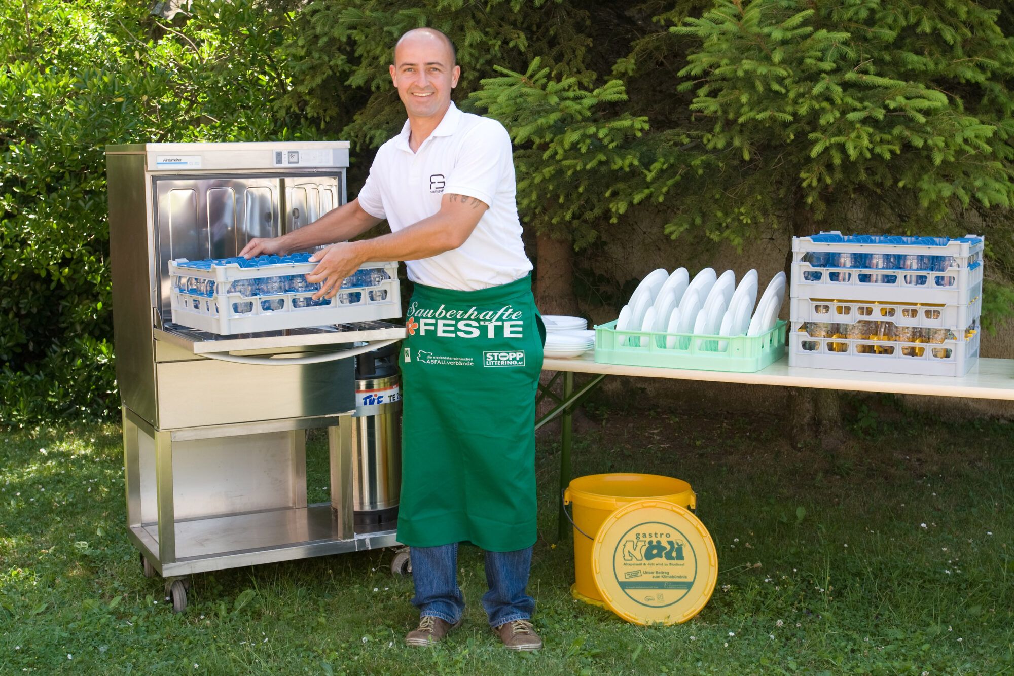 Ein Mann steht im Garten, er hat eine grüne Schürze um und räumt den Geschirrspüler des Festpaketes aus. Neben ihm am Tisch befinden sich saubere Teller und Gläser. Am Boden steht ein NÖLI Kübel.