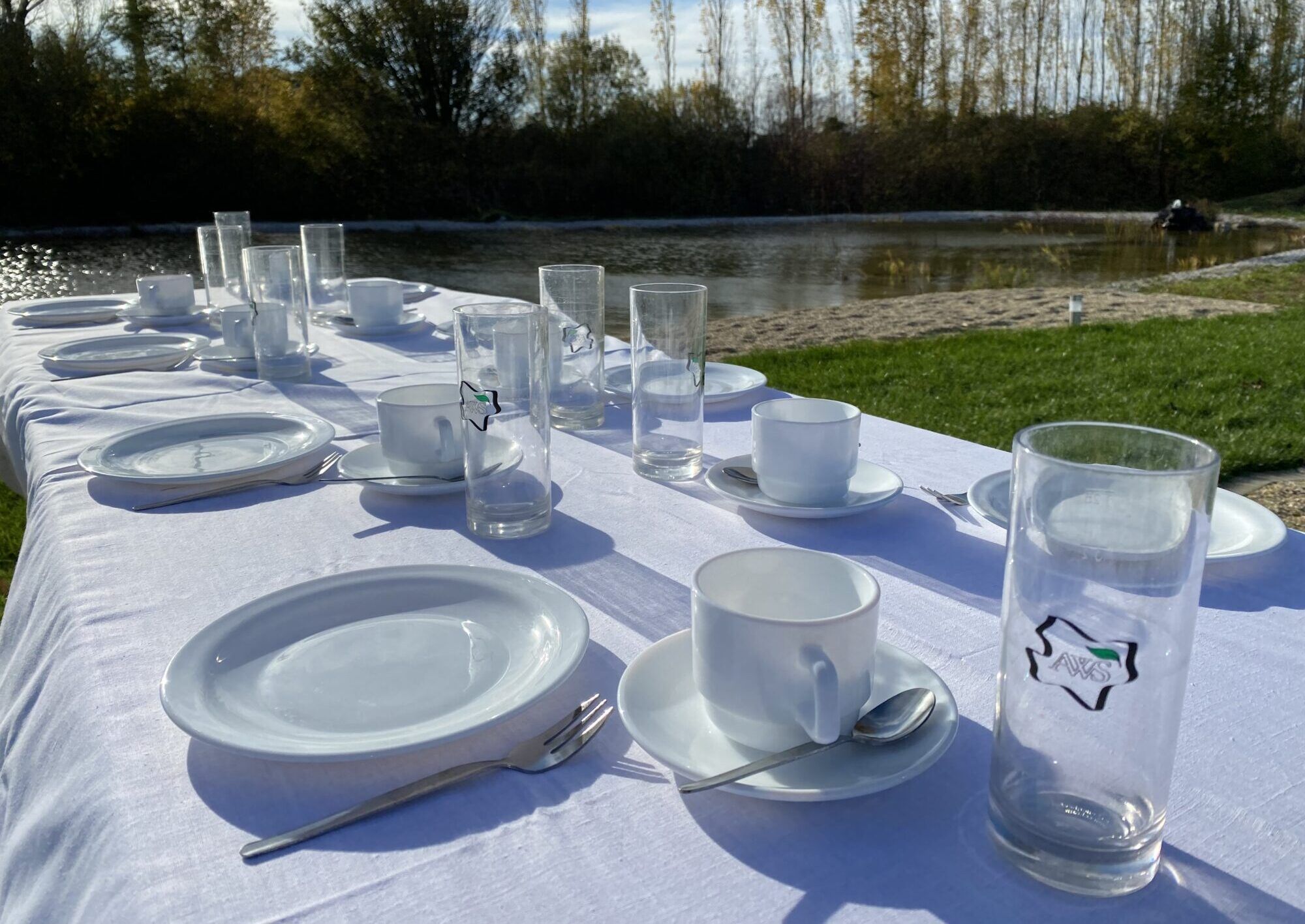 Gedeckter Tisch mit kleinem Teich und Bäumen im Hintergrund. Auf dem Tisch sieht man ein weißes Tischtuch, Gläser mit AWS Logo, Teller, Kuchengabeln und Kaffeetassen mit -untersetzern und -löffeln.