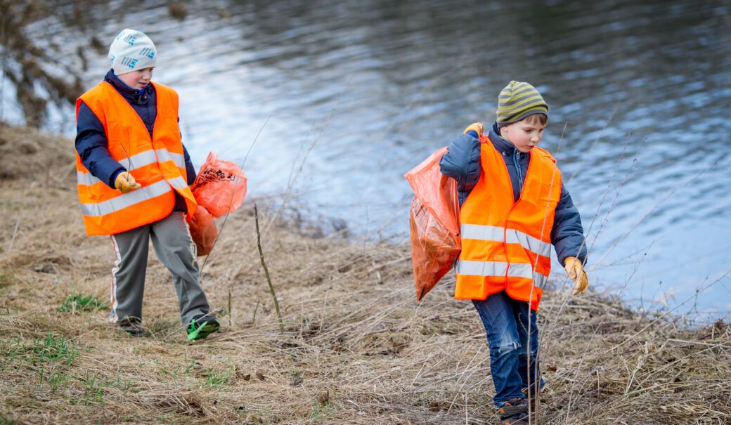 Auf einer Wiese neben einem Fluss sind zwei Buben unterwegs. Sie tragen Warnwesten und halten jeweils einen orangenen Sack in der Hand, während sie aufmerksam die Umgebung betrachten.