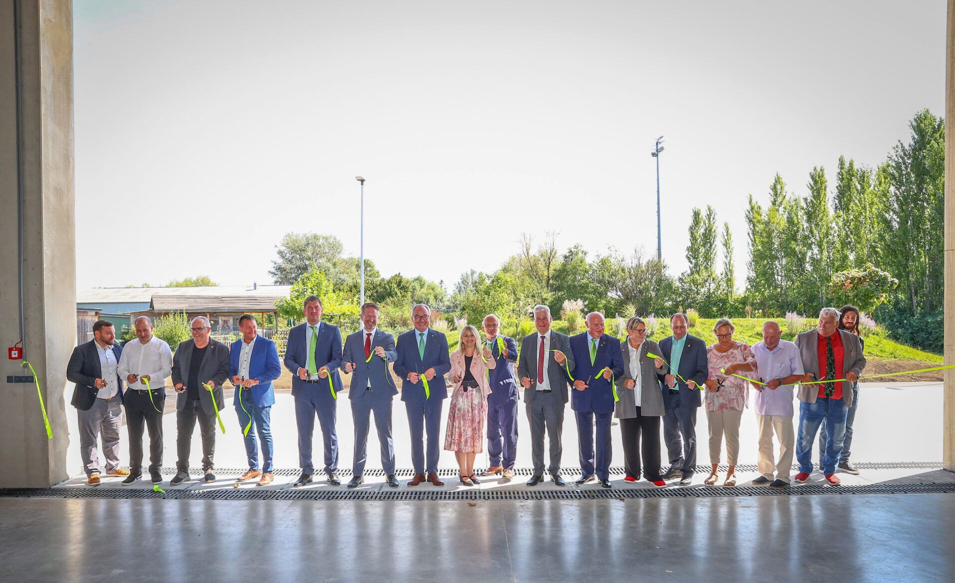 Band-Durchschneiden beim AWS Neubau. 17 Personen stehen mit Scheren an einem gr&uuml;nen Band und durchschneiden dieses feierlich als Akt der Er&ouml;ffnung.