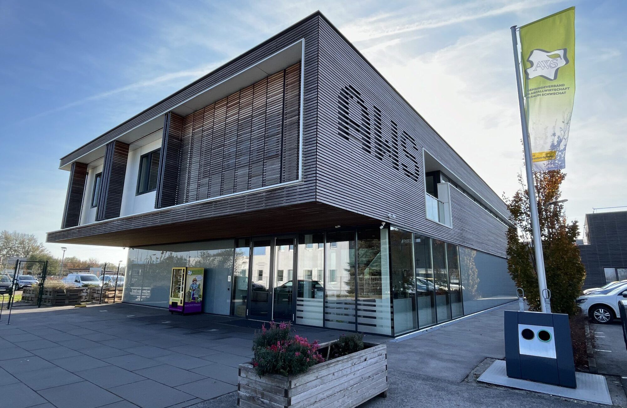 Das AWS Bürogebäude ist zu sehen, davor steht ein kleines Hochbeet, rechts ist eine grüne Fahne mit dem AWS Logo zu sehen und im Hintergrund ist der blaue Himmel zu sehen.