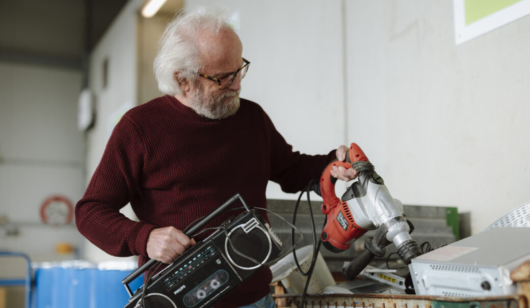 Ein älterer Herr mit Brille, einem weinroten Pullover und einer Jeans hält einen Radio und einen Schlagbohrer in der Hand, um diese in den vorhergesehenen Behälter der Sammelstelle zu entsorgen.