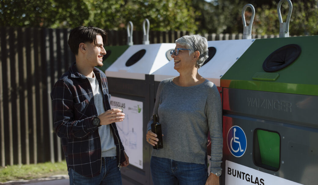 Eine ältere Dame mit grauen Haaren entsorgt bei einem Buntglas Container auf einer Sammelinsel ein dunkles Glas. Neben der Dame steht ein junger Mann mit Jean, Pullover und Weste und unterhält sich mit der Dame. Er hält ein weißes Glas in der Hand.