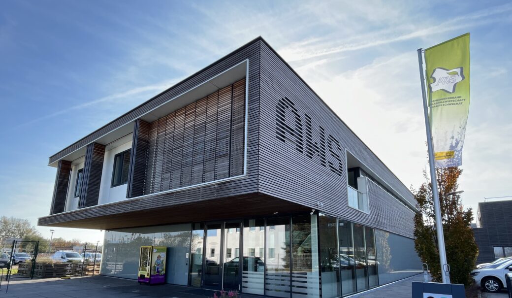 Das AWS Bürogebäude ist zu sehen, davor steht ein kleines Hochbeet, rechts ist eine grüne Fahne mit dem AWS Logo zu sehen und im Hintergrund ist der blaue Himmel zu sehen.