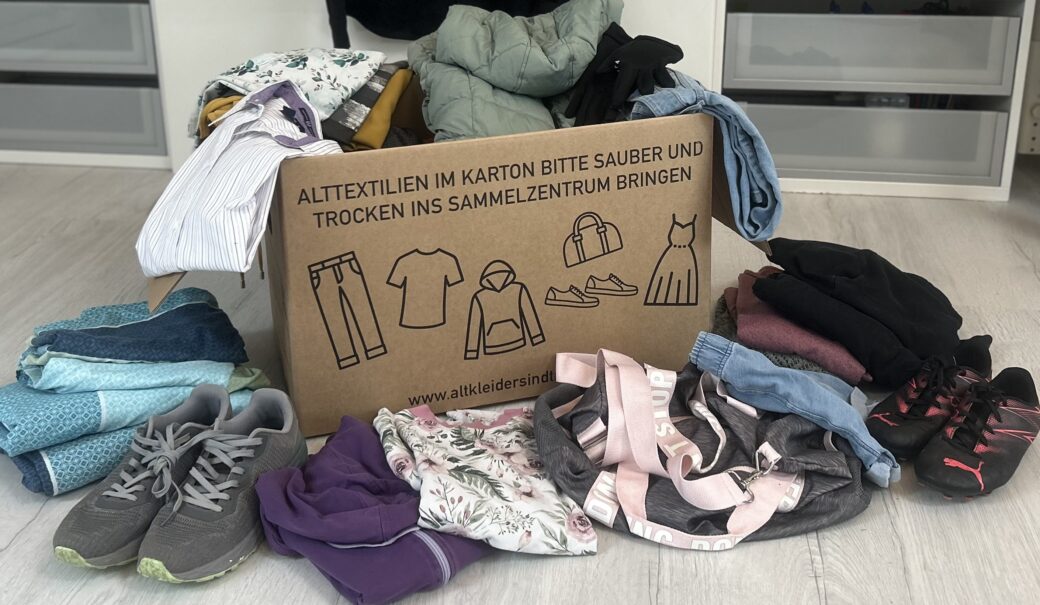 diverse Altkleider, Alttextilien und ein Paar SChueh, die in und rund um einen Karton liegen, auf dem schemenhaft Kleider und Schuhe abgedruckt sind