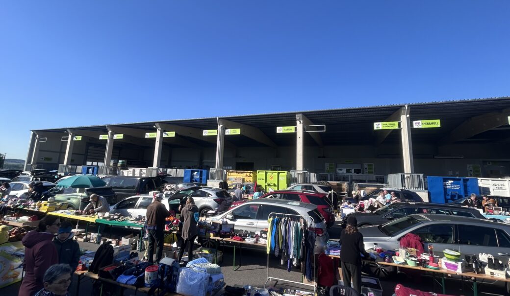 Flohmarkt am WSZ Fischamend. Im Vordergrund Verkaufsstände, Menschen, Autos. Im Hintergrund das Sammelzentrum mit Schildern und Containern.