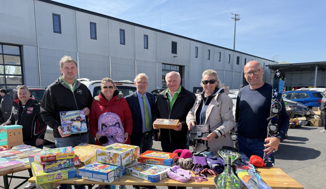 Personen stehen hinter einem Flohmarkt-Verkaufsstand und präsentieren Kinderspiele, Kinderski und einen Rucksack, auf dem Tisch liegen ganz viele Kinderspiele