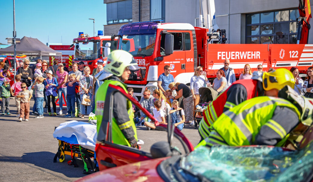 AWS Zivilschutztag 2026 Außengelände des Abfallverband Schwechat, blauer Himmel, im Vordergrund eine Einsatzvorführung mit einem inszenierten Autounfall mit Einsatzpersonal der Feuerwehr, im Hintergrund Feuerwehr Fahrzeuge und Zuschauerinnen und Zuschauer