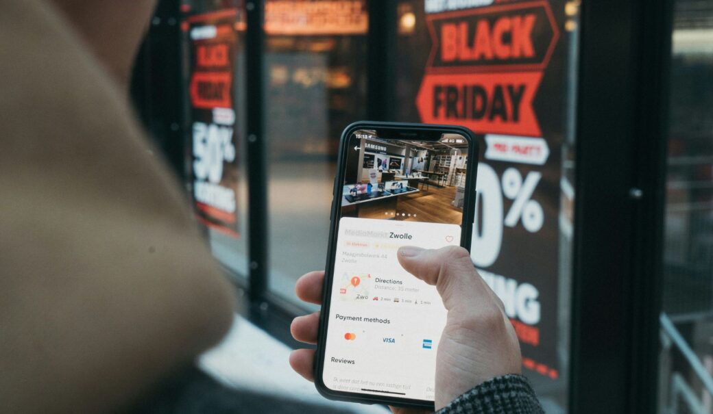 Mann steht vor Elektronikladen auf dem fett Black Friday aufgedruckt ist. W&auml;hrenddessen vergleicht er Preise auf seinem Handy